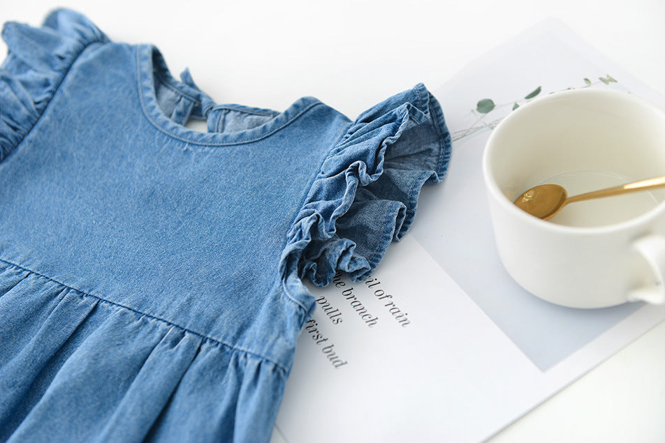 Blue girls denim dress with ruffled details on a white surface with a cup and spoon.