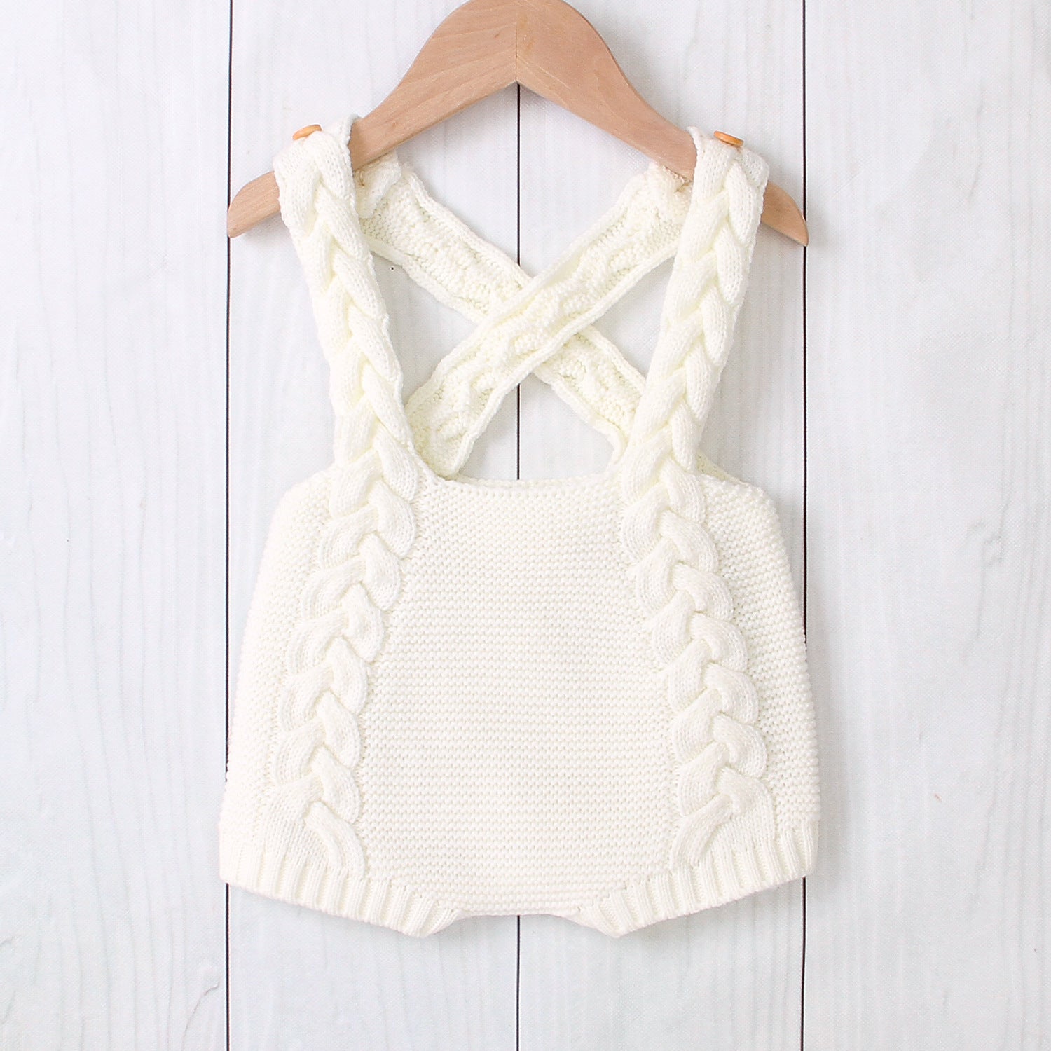 Light beige knitted romper with criss-cross back on a wooden hanger against a white wooden background